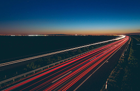 Autostrada o zmierzchu sfotografowana z długim czasem naświetlania; rozmyte światła jadących samochodów tworzą kolorowe smugi na tle ciemniejącego nieba.
