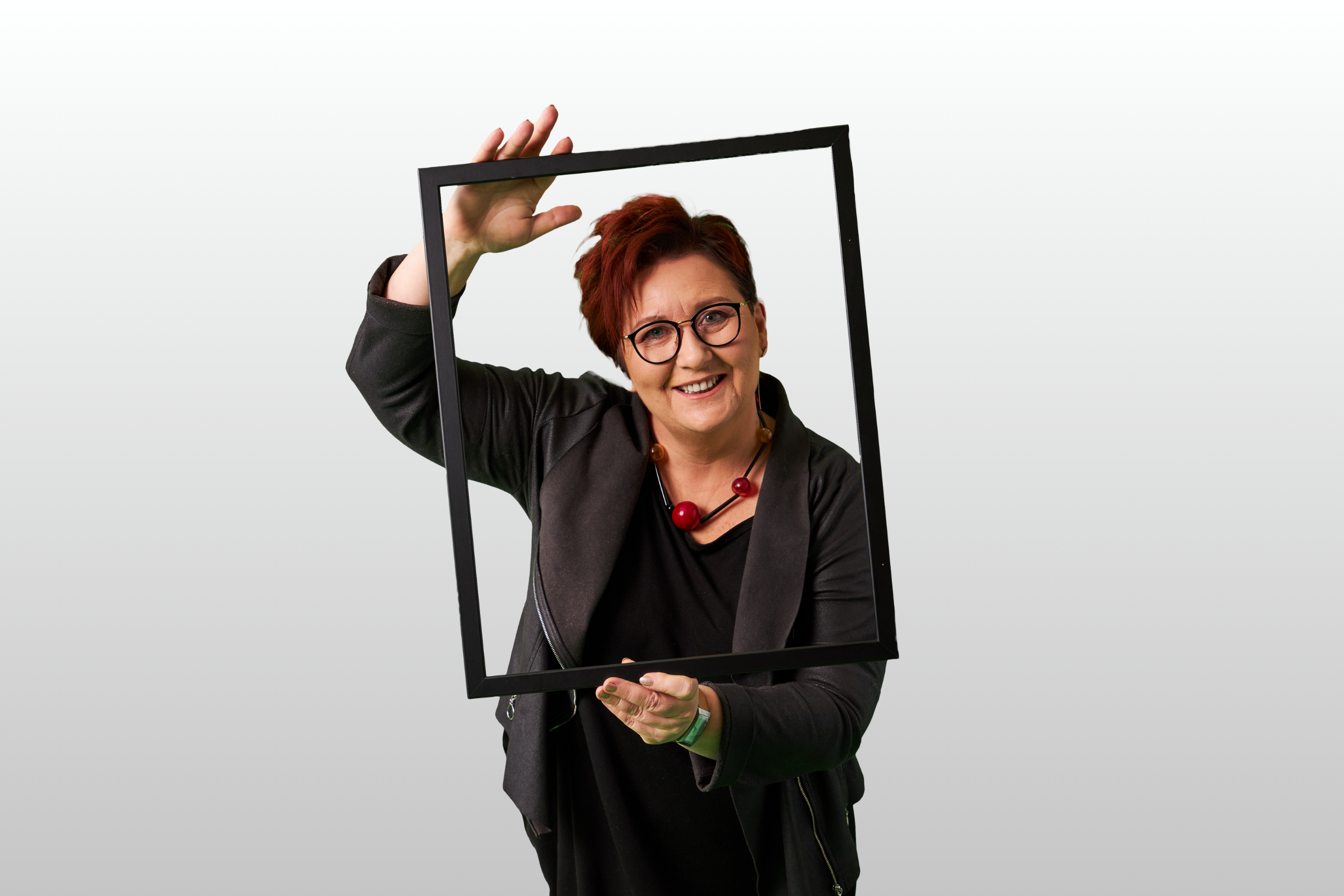 Uśmiechnięta kobieta z krótkimi rudymi włosami i okularami trzyma przed sobą czarną ramkę na zdjęcia, żartobliwie pozując na prostym jasnoszarym tle. Ma na sobie ciemną kurtkę i masywny czerwony naszyjnik.