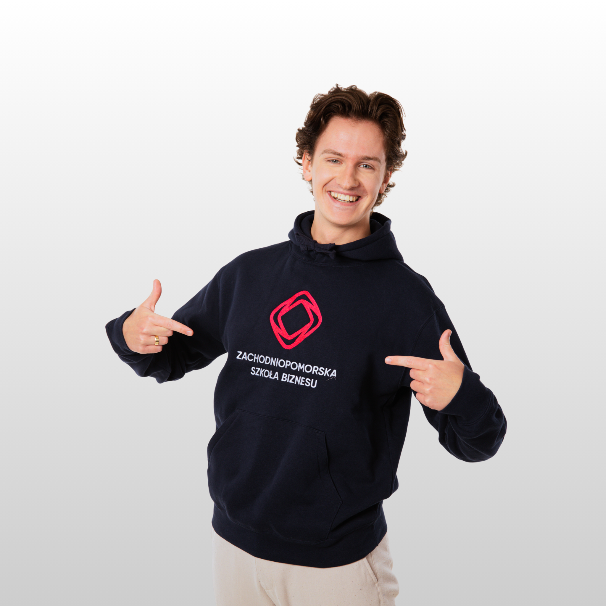 Młody, uśmiechnięty mężczyzna w granatowej bluzie, stoi na jasnym tle i obiema rękami wskazuje logo Zachodniopomorskiej Szkoły Biznesu, które się znajduje na przodzie bluzy. 