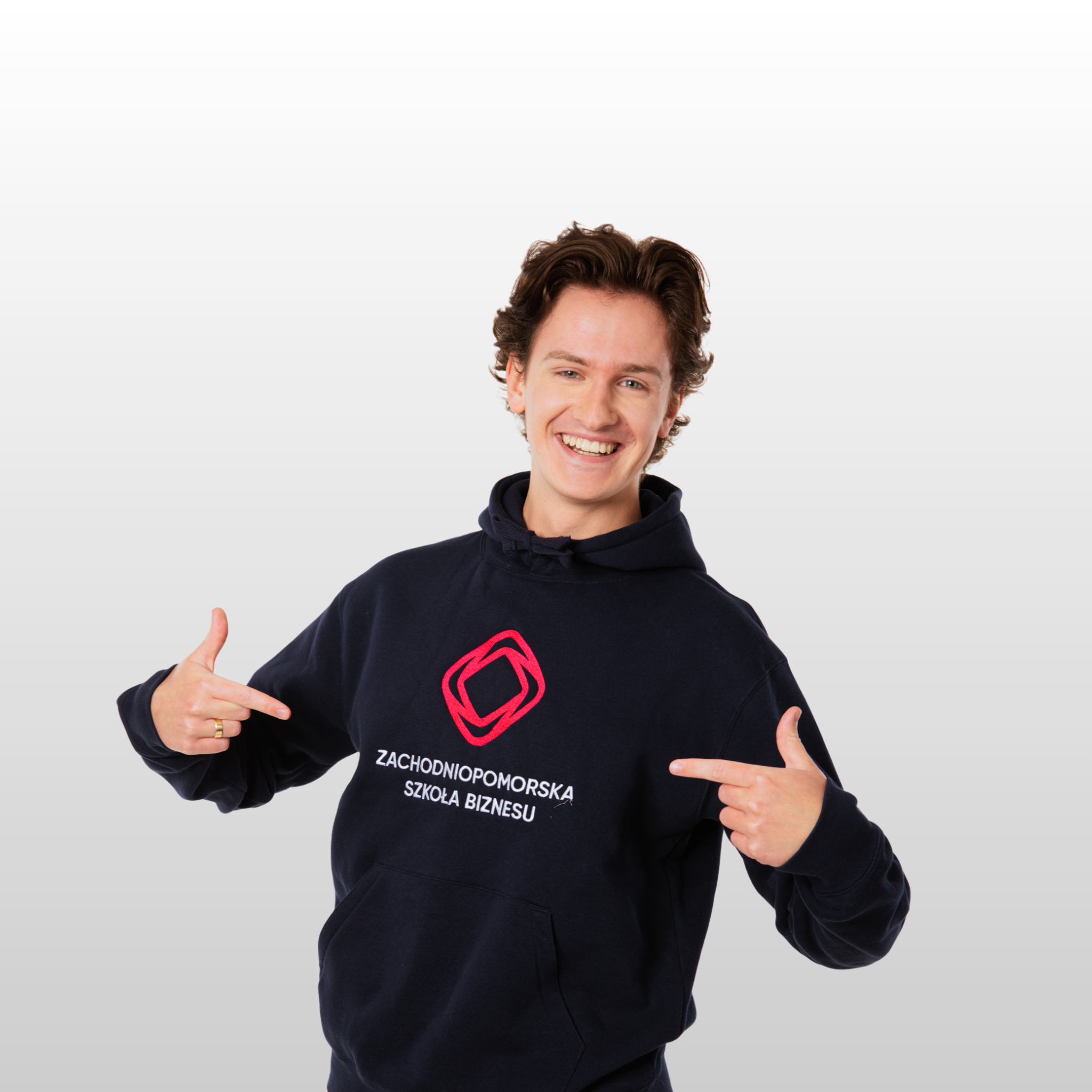 Młody mężczyzna uśmiecha się i wskazuje na logo i tekst na swojej granatowej bluzie z kapturem, który brzmi Zachodniopomorska Szkoła Biznesu. Stoi na prostym, jasnym tle.