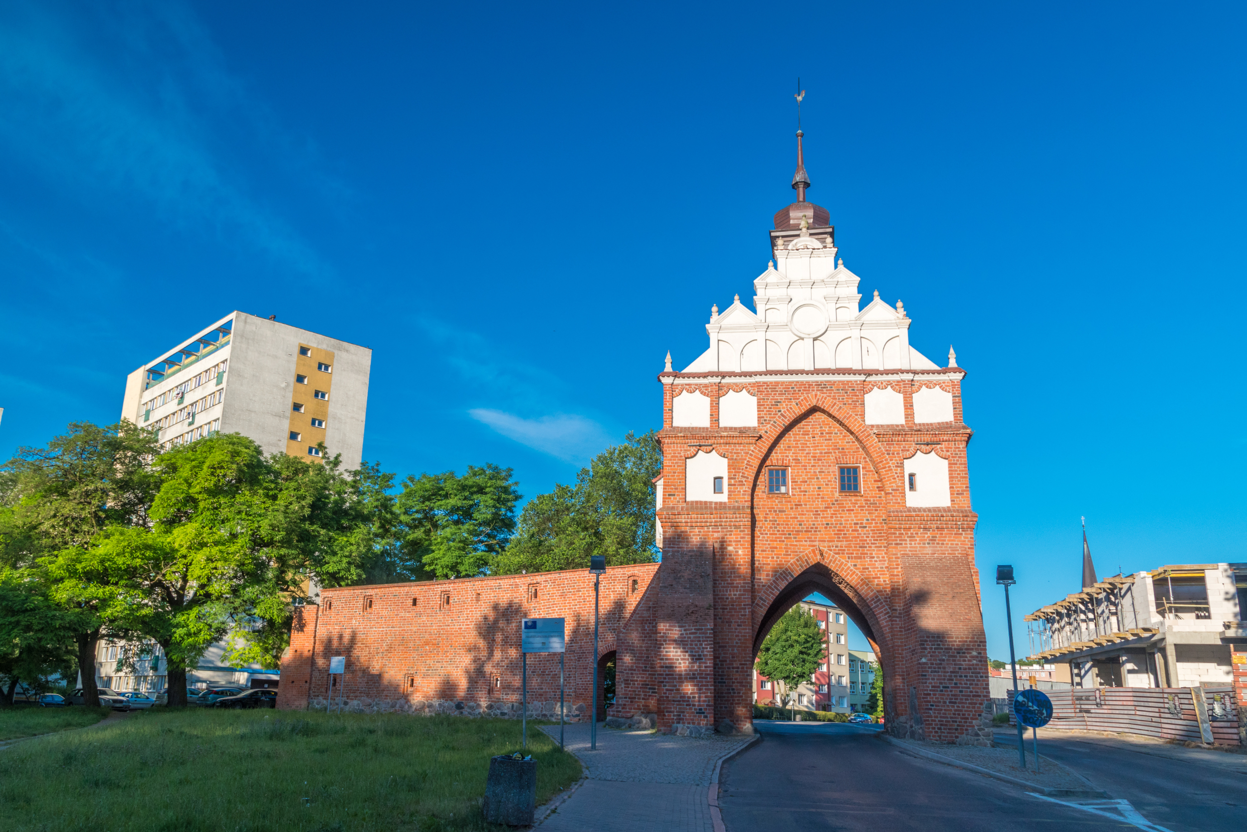 Zabytkowa ceglana brama miejska z białą iglicą stoi przed czystym błękitnym niebem, otoczona nowoczesnymi budynkami, drzewami i brukowaną drogą prowadzącą przez łuk.
