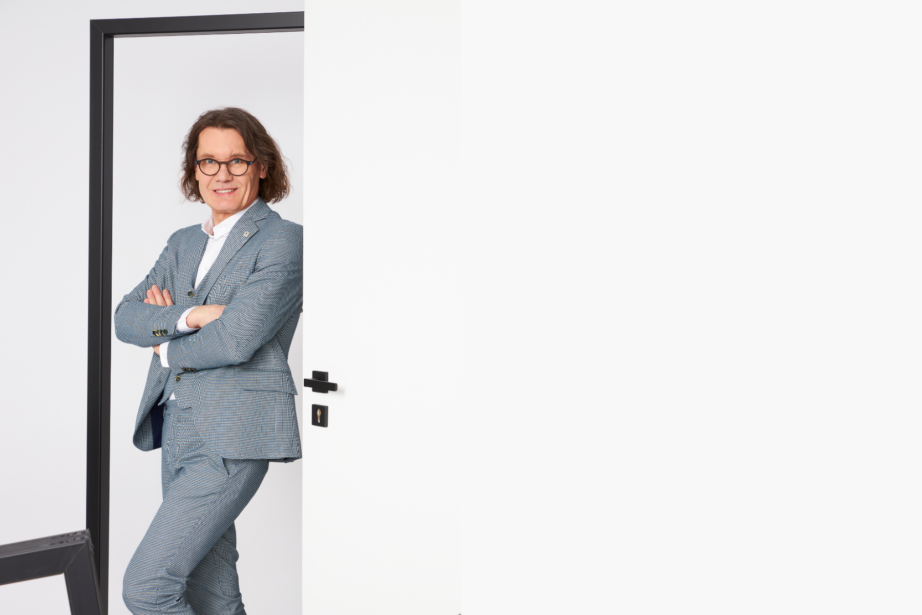 Po lewej stronie stoi elegancki mężczyzna w okularach szarej marynarce, opiera się o framugę drzwi