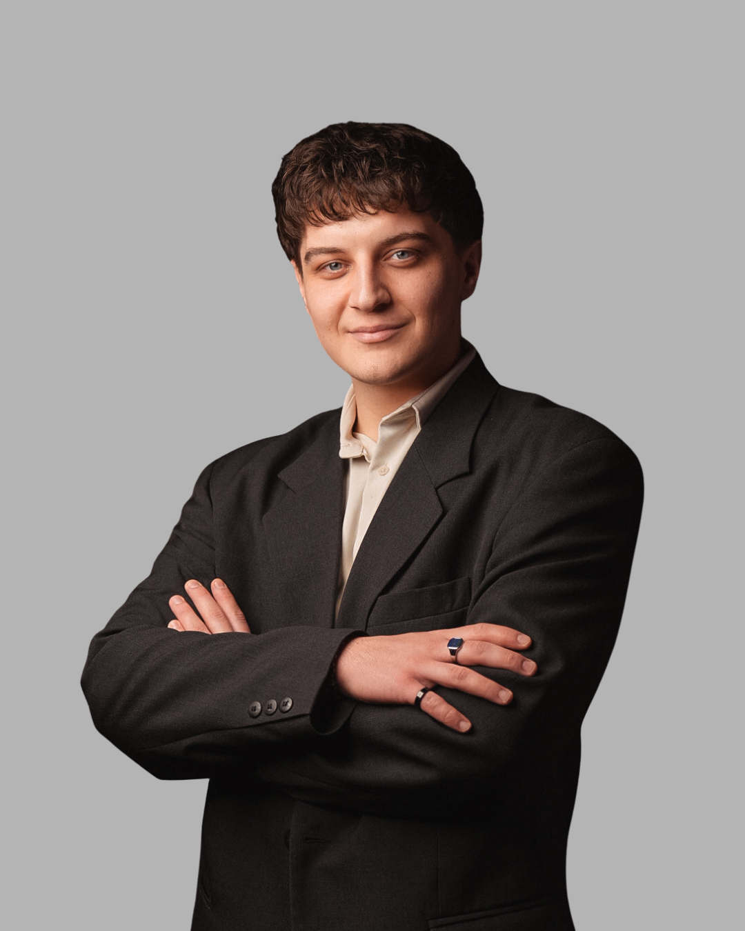 Rekruter studentów w ciemnej marynarce i jasnej koszuli, stojący z założonymi rękami, patrząc w obiektyw na neutralnym, jasnoszarym tle.