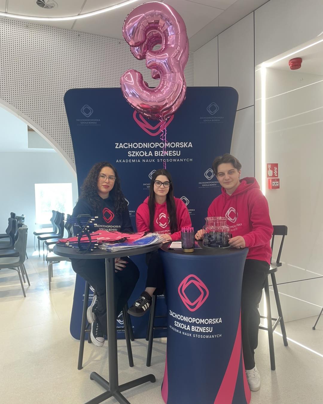 Grupa trzech studentów w bluzach ZPSB na wydarzeniu, siedzą na wysokich krzesłach przy trybunce i ściance z logo uczelni