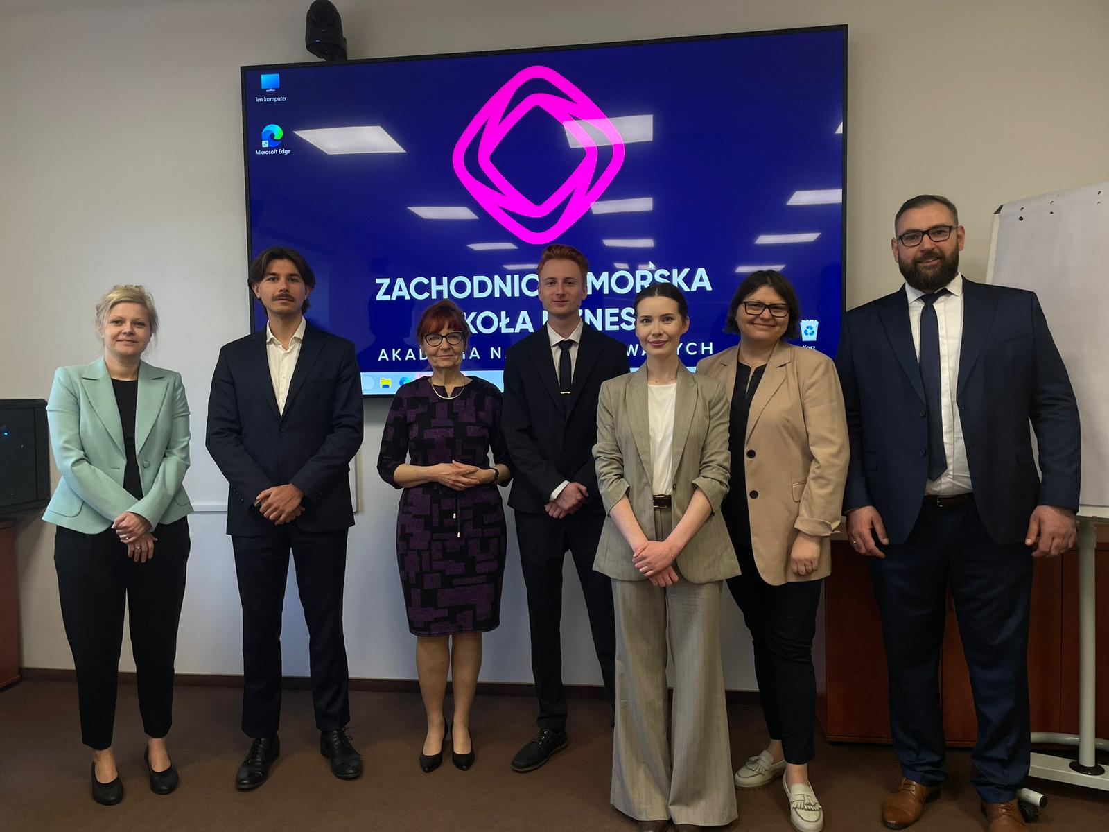 Grupa ludzi elegancko ubranych pozuje na tle ekranu z logo uczelni