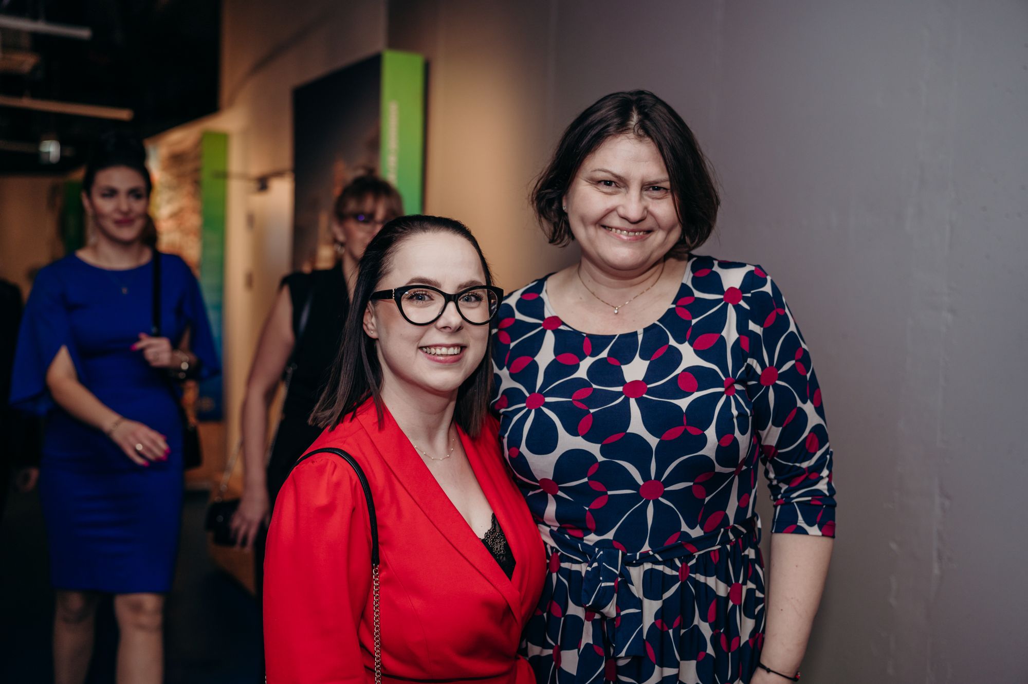 Dwie uśmiechnięte kobiety pozują do zdjęcia w eleganckim wnętrzu; jedna w czerwonej marynarce i okularach, druga w sukience w geometryczny wzór, w tle inne uczestniczki wydarzenia.
