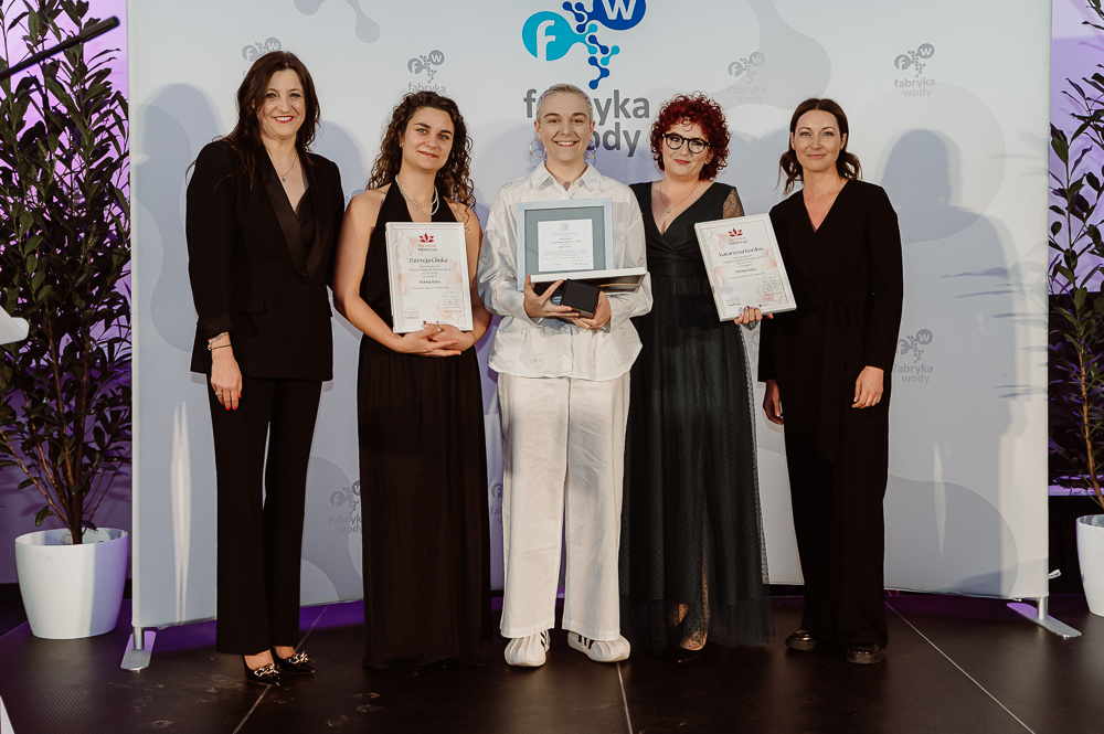 Pięć osób stoi na scenie podczas wręczenia nagród; trzy osoby trzymają dyplomy i statuetki, wszyscy ubrani elegancko na tle ścianki z logo „fabryka wody”.