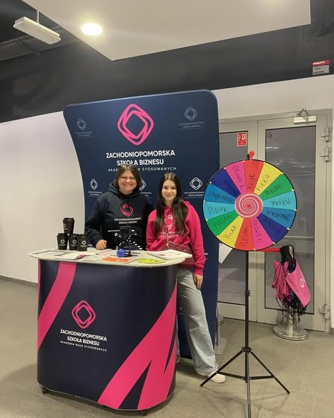 Dwie kobiety na stoisku reklamowym uczelni pozują do zdjęcia