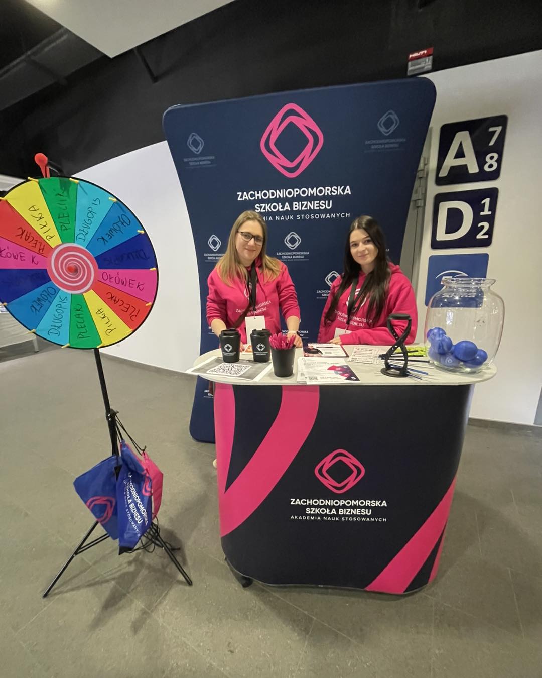 Dwie kobiety na stoisku reklamowym uczelni pozują do zdjęcia