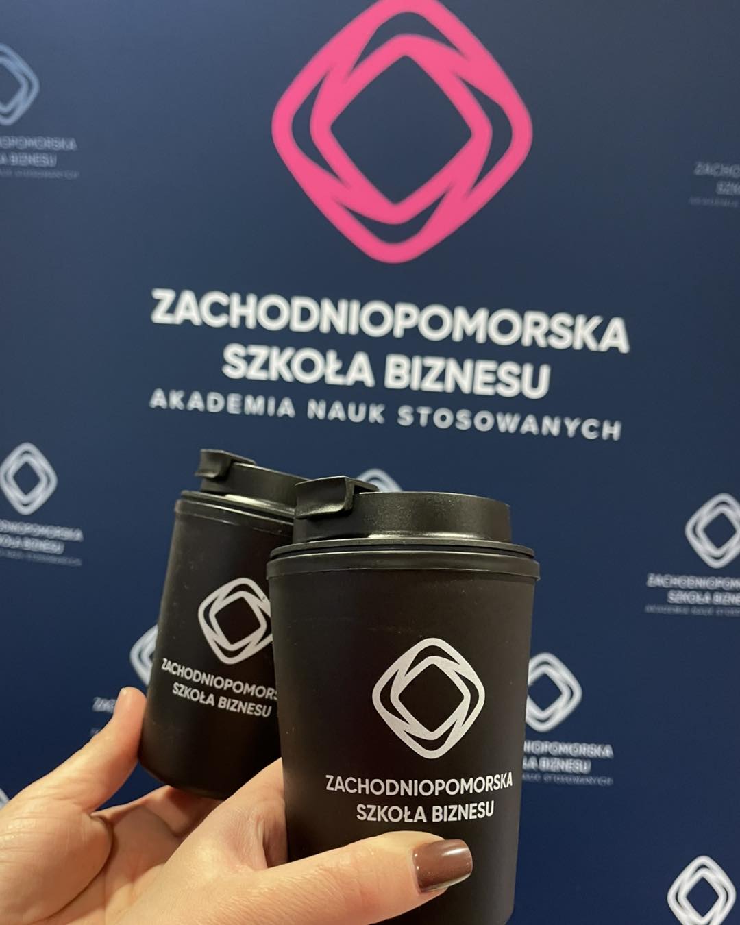 Dwa kubki plastikowe z logo zachodniopomorskiej szkoły biznesu prezentowane na tle ścianki reklamowej