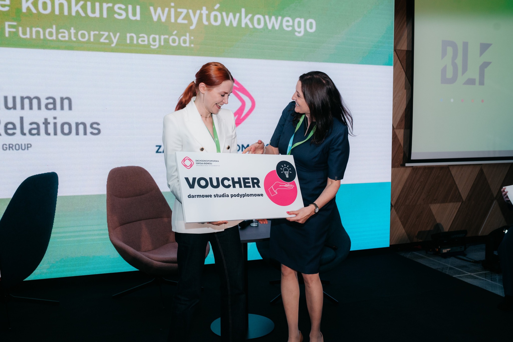 Rektor przekazuje duży voucher na scenie zwyciężczyni losowania. Obie uśmiechają się.