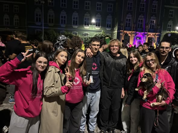 Grupa młodych osób stoi razem na tle tłumu podczas wieczornego wydarzenia pod Urzędem Miasta w Szczecinie. Kilka osób ma różowe bluzy z logo,