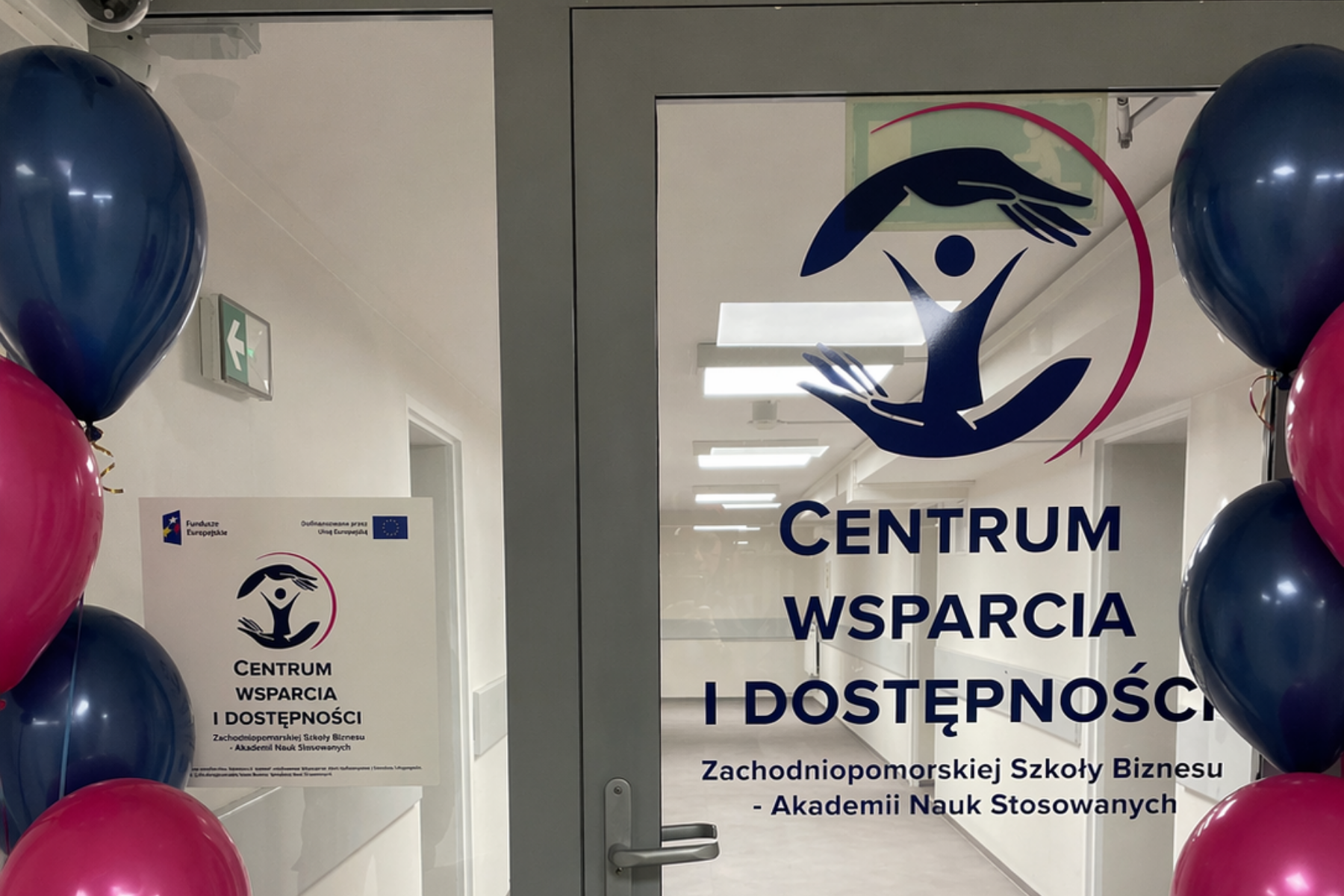 Logo centrum wsparcia i dostępności na szklanych drzwiach z szarą ramą po bokach widać granatowe i różowe balony
