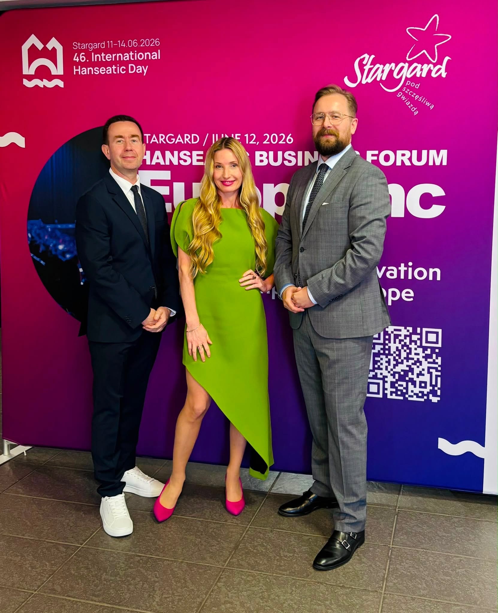 Trzy osoby pozują na tle fioletowej ścianki reklamowej z logo Stargardu i napisem „HANSEATIC BUSINESS FORUM”. W środku stoi kobieta w limonkowej sukience i różowych szpilkach, po jej lewej stronie mężczyzna w ciemnym garniturze i białych butach, a po prawej mężczyzna w szarym garniturze i okularach.