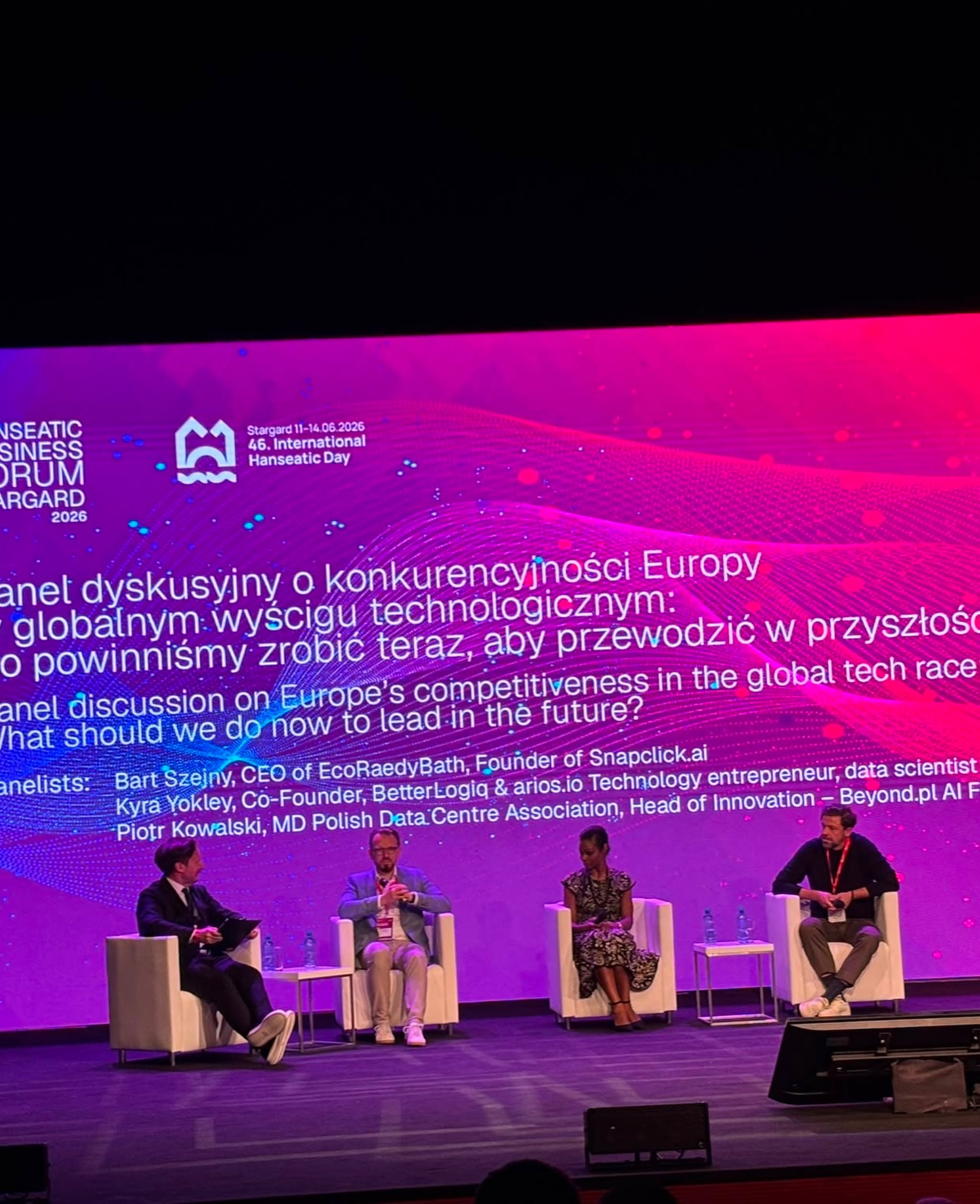 Panel dyskusyjny na scenie. Cztery osoby siedzą w białych fotelach przed wielkim różowym ekranem z napisem „Panel dyskusyjny o konkurencyjności Europy w globalnym wyścigu technologicznym...”. Na ekranie widoczne są także nazwiska prelegentów.
