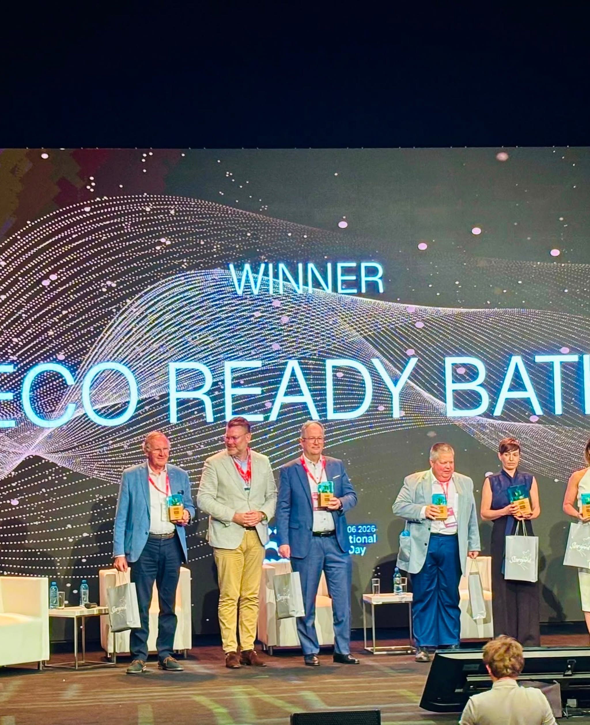Grupa pięciu laureatów stoi na scenie podczas gali, trzymając w dłoniach statuetki i upominki. W tle widoczny jest wielki ekran z grafiką fal i napisem „WINNER ECO READY BATH”. Na dole kadru widoczny jest tył głowy jednego z uczestników na widowni.