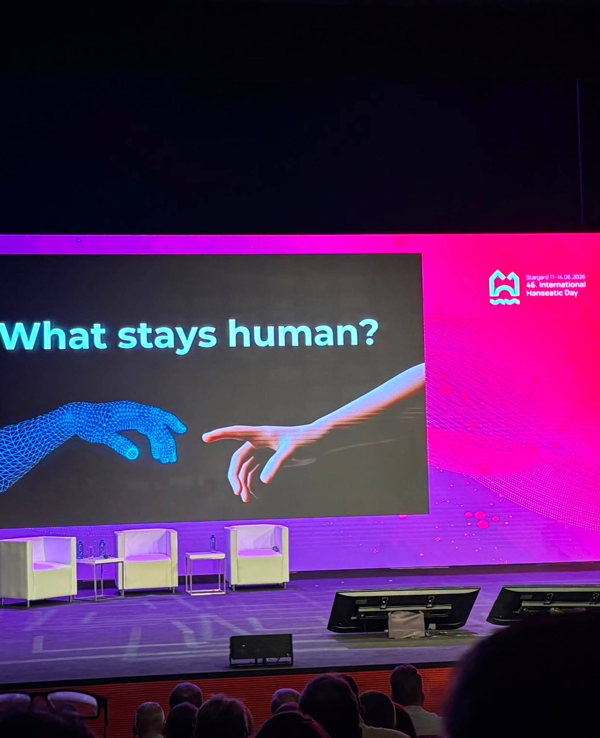 Duży ekran sceniczny z napisem „What stays human?”. Pod tekstem grafika przedstawiająca zbliżające się do siebie dłonie: po lewej cyfrowa, niebieska sieć 3D, po prawej ludzka dłoń. W prawym górnym rogu logo 46. International Hanseatic Day Stargard 2026. Przed ekranem pusta scena z białymi fotelami.